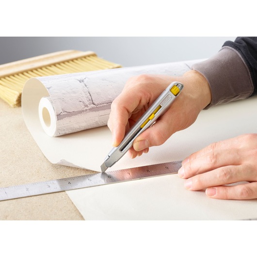 Photo of STANLEY 0-10-095 utility knife cutting wallpaper along a metal ruler, with wallpaper roll and brush visible.