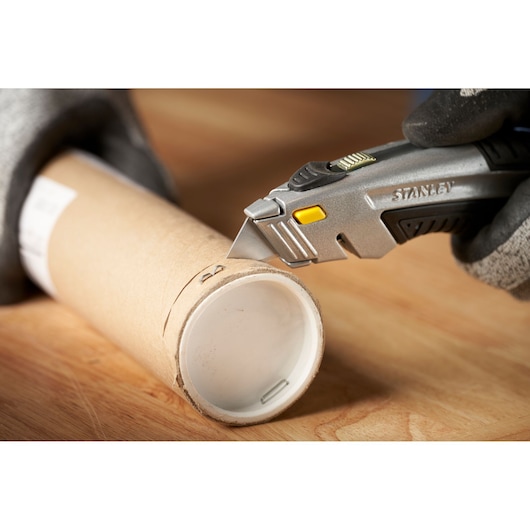 Photo of STANLEY 0-10-788 utility knife cutting cardboard tube on wood surface, close-up side angle.