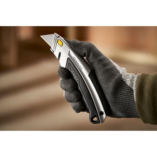 Photo of STANLEY 0-10-788 utility knife held in gloved hand, silver body with black grip and yellow button.