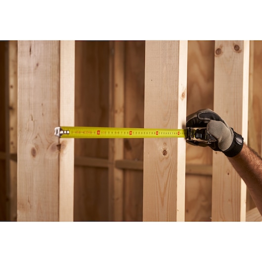STANLEY 0-33-552 tape measure in use, measuring between wooden beams, held by gloved hand.