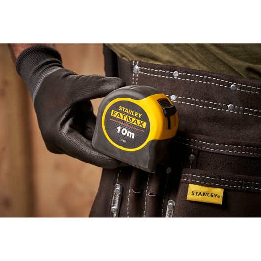 STANLEY 0-33-811 tape measure FATMAX 10m held by gloved hand, clipped to tool belt, close-up view.