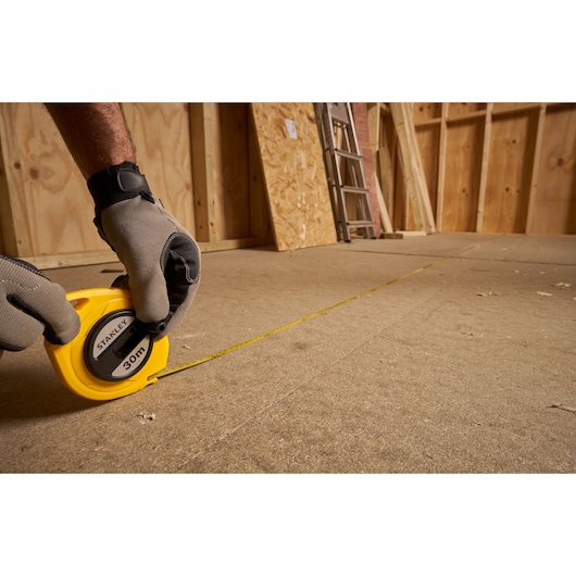 STANLEY 0-34-108 measuring tape, yellow, used on construction floor, person wearing gloves.