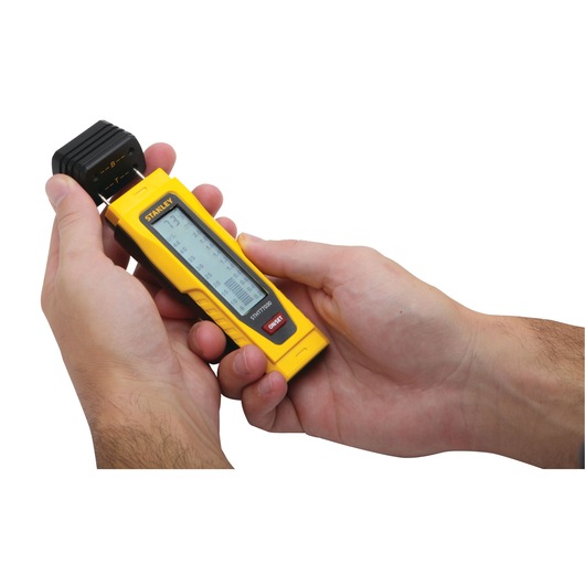 Photo of STANLEY 0-77-030 digital moisture meter handheld device in use, yellow and black body, close-up view.