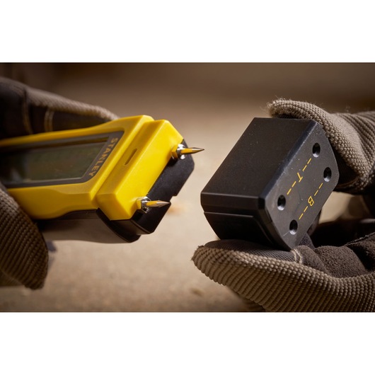 STANLEY 0-77-030 digital moisture meter close-up with probe points and calibration block held by gloved hands.