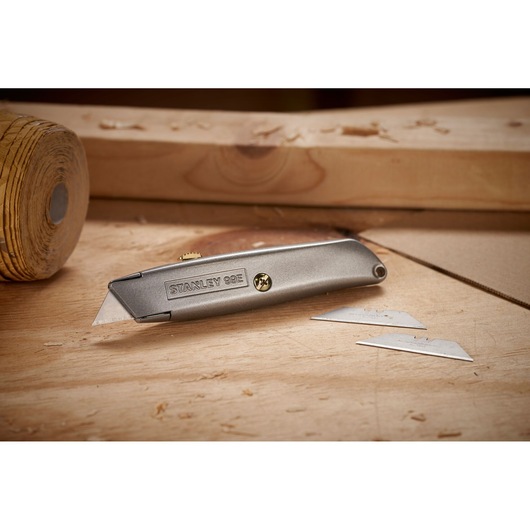 A metal Stanley 99E utility knife placed on a wooden surface, with two spare blades lying nearby. The background shows woodworking materials and tools.