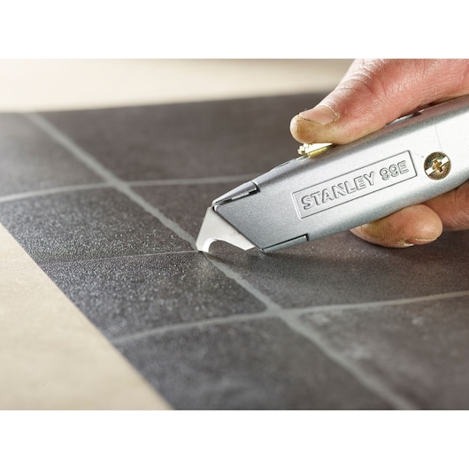 Photo of STANLEY 2-11-983 utility knife scoring black tile, close-up of blade and hand.