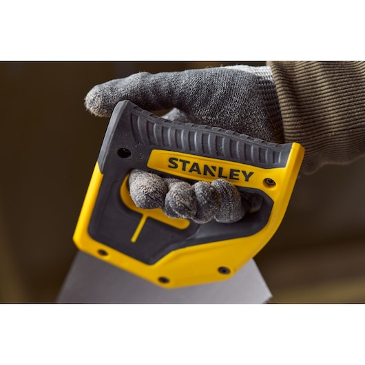 Close-up of a gloved hand holding a STANLEY 2-15-244 hand saw with a yellow and black ergonomic handle.