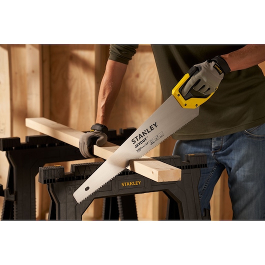 Stanley 2-15-289 hand saw with yellow handle cutting wood on black sawhorse, workshop background.