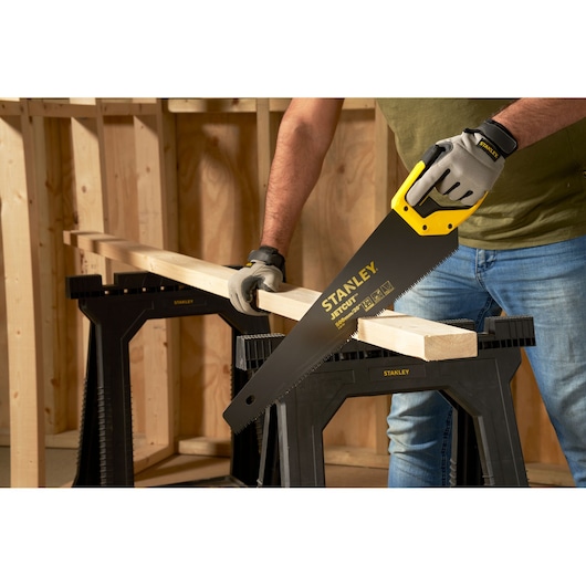 Photo of STANLEY 2-20-151 hand saw cutting wood plank, person wearing gloves, wood sawhorses in workshop.