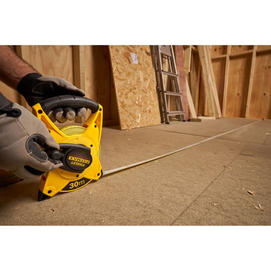 STANLEY 2-34-814 measuring tape, 30m, on building site floor with gloves holding handle, side angle.