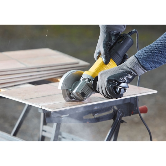 STANLEY FMEG210 angle grinder cutting tile, black and yellow body, user with gloves, close-up action shot.