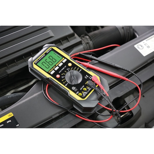 STANLEY FMHT0-77419 digital multimeter with yellow accents, shown on a car engine during electrical testing.