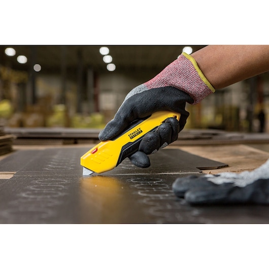 Stanley FMHT10369-0 utility knife cutting material, held by gloved hand in industrial setting.