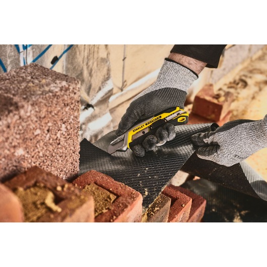 STANLEY FMHT10594-0 utility knife cutting black material near bricks, close-up photo.