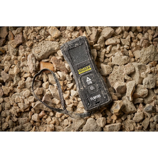 STANLEY FMHT77165-0 laser distance measurer with strap on rocky ground, covered in dust and water drops.
