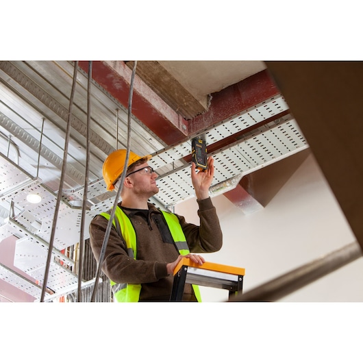 Photography of STANLEY FMHT77652-0 digital detector used by worker in safety gear on ladder at construction site.