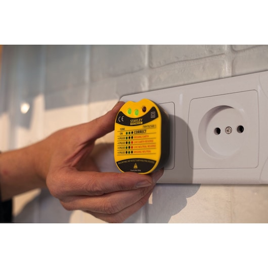 Photo of STANLEY FMHT82569-6 socket tester plugged into white wall outlet, yellow and black device held by hand.