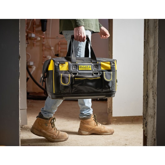 STANLEY FMST1-71180 tool bag, black and yellow, carried by person in work boots, indoor setting.