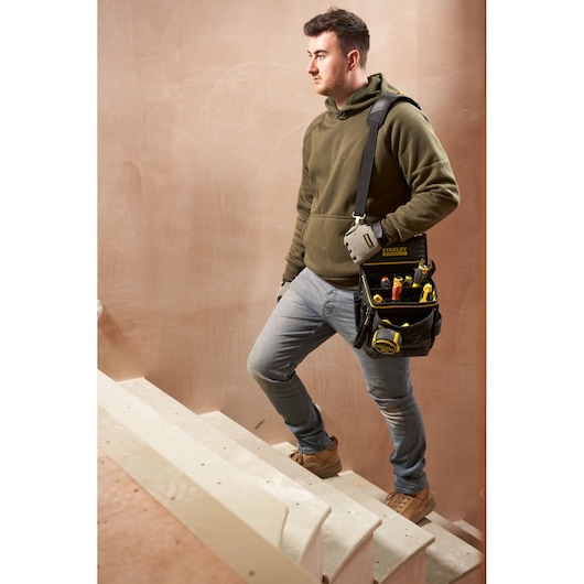 Photo of person carrying STANLEY FMST17624-1 tool bag with tools over shoulder, standing on stairs at a construction site.