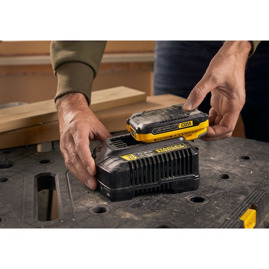 Photo of STANLEY SFMCB14 battery charger with battery on workbench, hands placing battery onto charger.