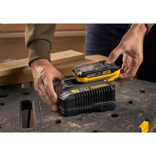 STANLEY SFMCB202 battery being placed on black charger, close-up with workbench in background.