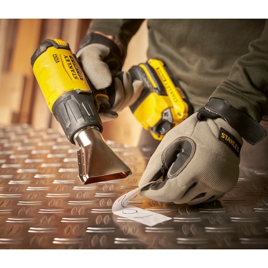 STANLEY SFMCE530B heat gun in use on metal sheet, worker wearing gloves, close-up angle.