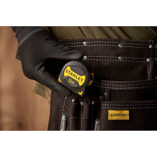 Stanley STHT0-33559 tape measure held in black-gloved hand clipped to brown tool belt.