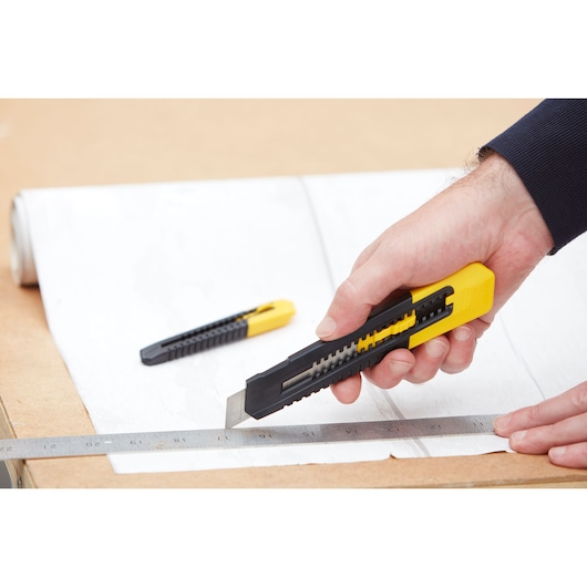 STANLEY STHT10202-0 utility knife with black and yellow handle cutting paper along a ruler, second knife in background.
