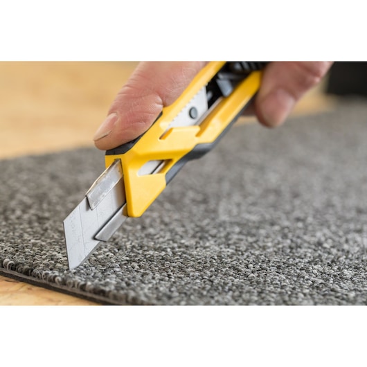 Close-up of a hand using a yellow utility knife to cut grey carpet.