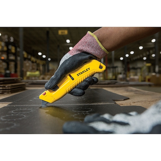 STANLEY STHT10364-0 utility knife cutting cardboard, worker wearing gloves, indoor warehouse setting.