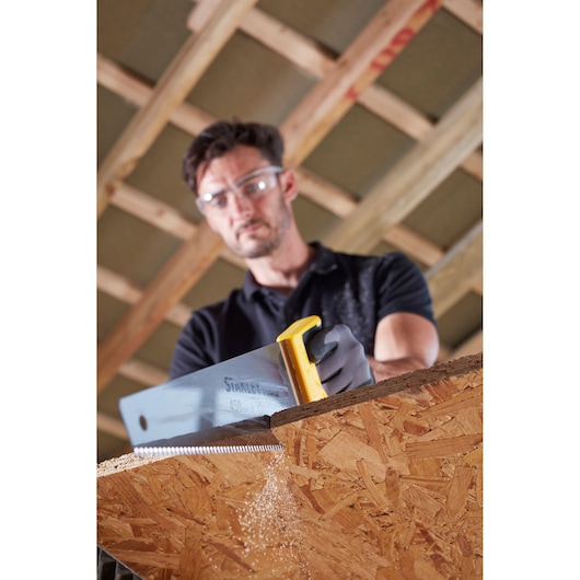 Photo of STANLEY STHT20354-1 handsaw cutting wood panel, close-up showing sawdust and yellow handle.