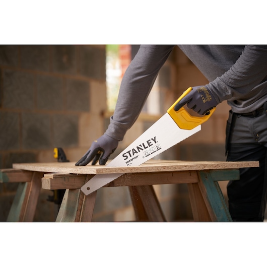 STANLEY STHT20367-1 hand saw with yellow handle cutting wood, side angle view, used by person wearing gloves.