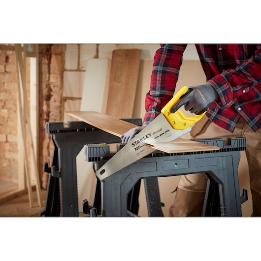Stanley STHT20369-1 hand saw with yellow handle cutting wood plank on black sawhorses in workshop.