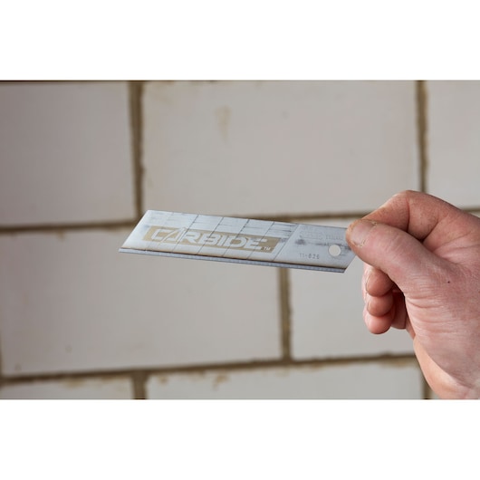 Photo of STANLEY STHT3-11825 utility blade held by hand, silver finish, tiled wall background.