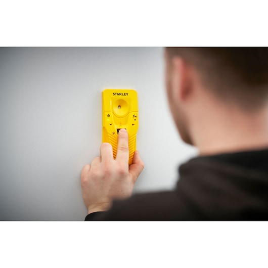 Photo of STANLEY STHT77587-0 yellow stud finder pressed against a white wall, held by a person.