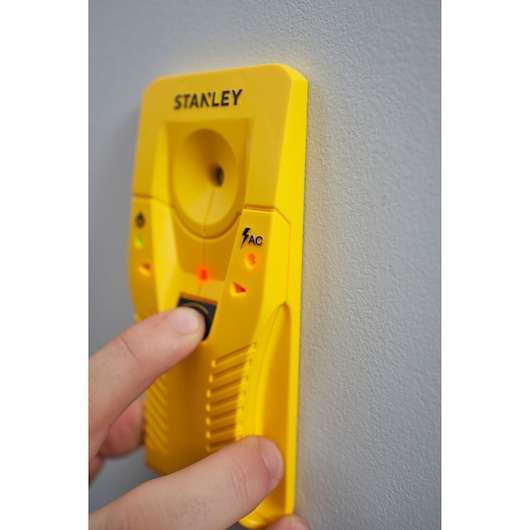 STANLEY STHT77587-0 yellow stud finder with AC detection held against a wall, close-up of hand pressing button.