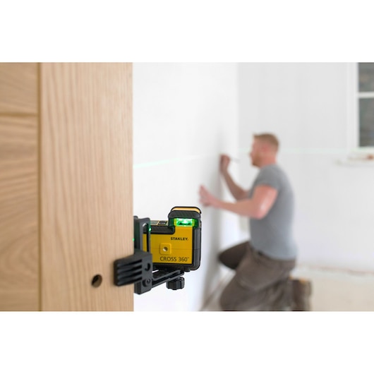 STANLEY STHT77594-1 cross line laser level mounted on wood, projecting green laser line, man working in background.