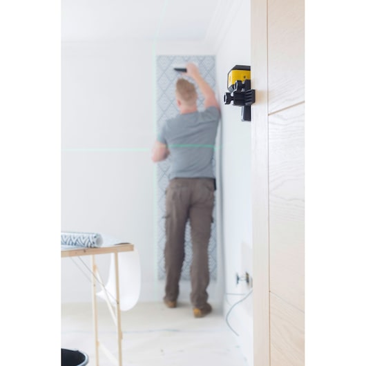 Photo of STANLEY STHT77649-1 laser level mounted on wall in home renovation scene, blurred person hanging wallpaper.