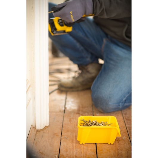 STANLEY STST82710-1 yellow storage bin with screws, used on wooden floor during construction, person in work gloves and boots visible.