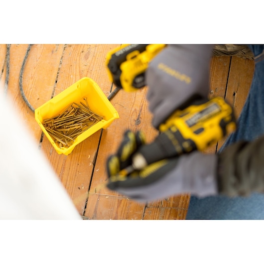 STANLEY STST82710-1 yellow bin with screws on wood floor, gloved hand holding drill.